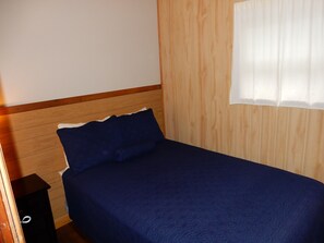 A single bed with a wooden headboard and a dark blue bedspread in a room with wooden paneling and a window with white curtains.