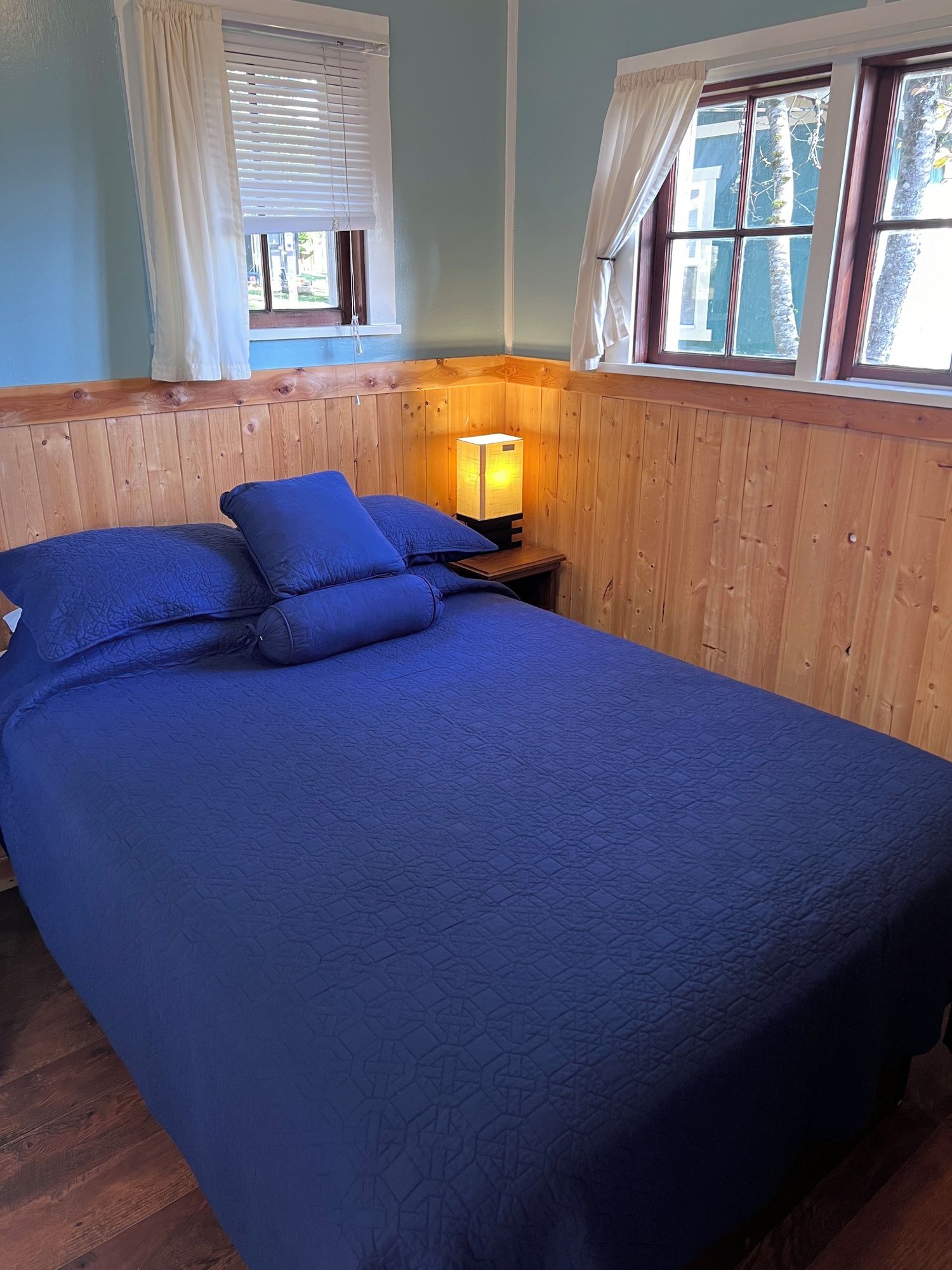 A room with a large bed, wooden walls, a nightstand with a lamp, and windows with curtains.