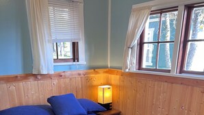 Una habitación con una cama grande, paredes de madera, una mesita de noche con lámpara y ventanas con cortinas.