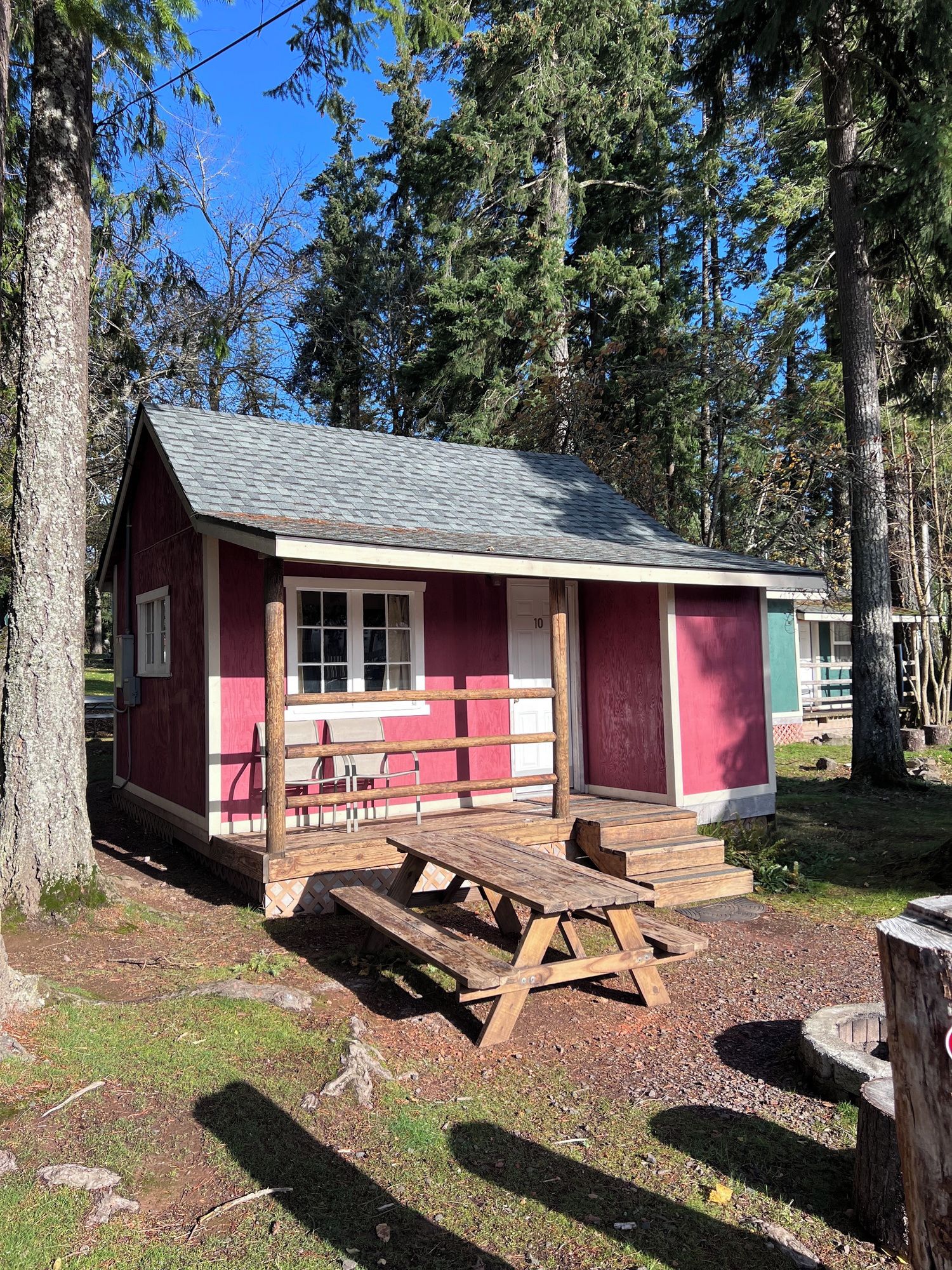 A small, single-story cabin with a red exterior and a grey roof, surrounded by trees.