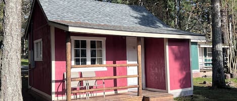 A small, single-story cabin with a red exterior and a grey roof, surrounded by trees.