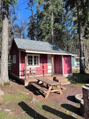 A small, single-story cabin with a red exterior and a grey roof, surrounded by trees. - Cabin 7: A Cozy Retreat Just a Short Walk from the Lake (Silver Lake)