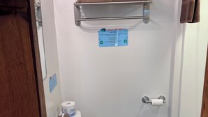 Un baño con inodoro, lavamanos y ducha. Hay dos toallas en un estante sobre el inodoro. Se ve un cartel en la pared.