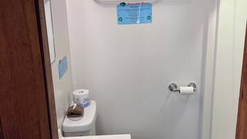 A bathroom with a toilet, sink, and shower. There are two towels on a shelf above the toilet. A sign is visible on the wall.