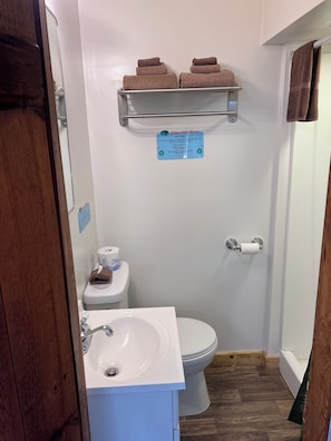 A bathroom with a toilet, sink, and shower. There are two towels on a shelf above the toilet. A sign is visible on the wall. - Cabin 7: A Cozy Retreat Just a Short Walk from the Lake (Silver Lake)
