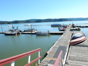 Un muelle de madera con barcas amarradas al final, cielo despejado y un lago tranquilo con una orilla boscosa al fondo.