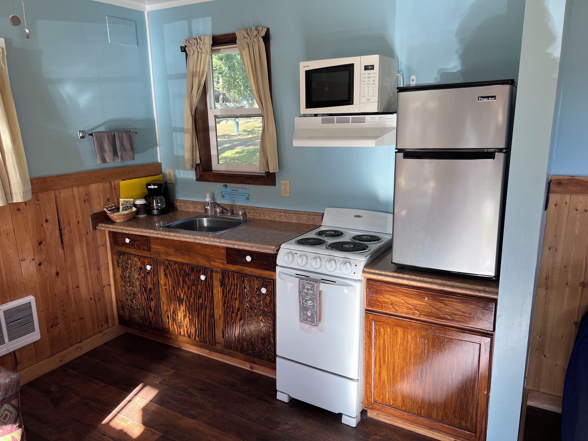 Una cocina compacta con estufa, microondas y refrigerador, armarios de madera y una ventana con cortinas.