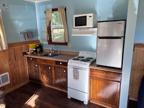 A compact kitchen with a stove, microwave, and refrigerator, wooden cabinets, and a window with curtains. - Cabin 7: A Cozy Retreat Just a Short Walk from the Lake (Silver Lake)