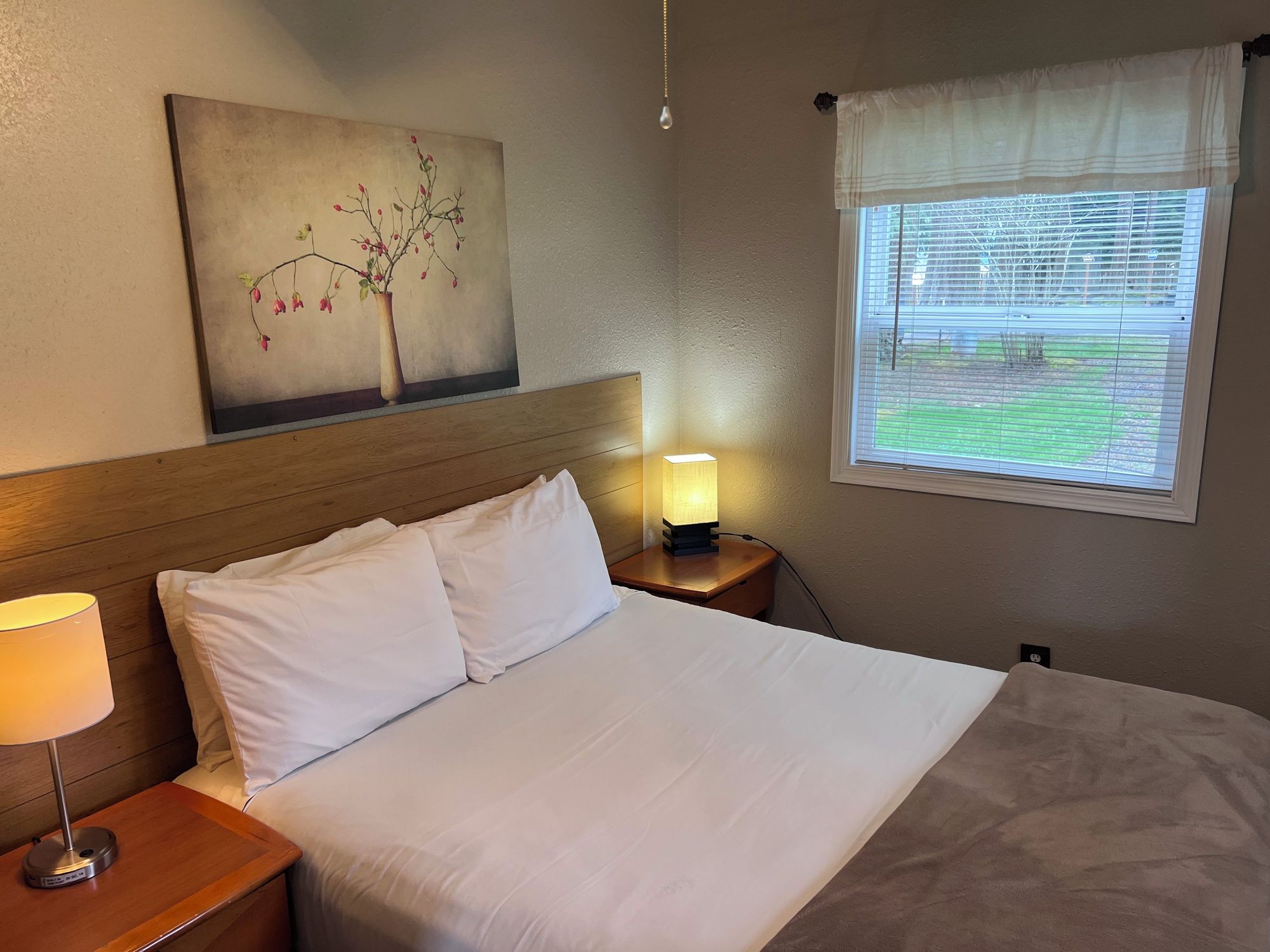 Een hotelkamer met een bed, een bedlamp, een nachtkastje, een schilderij aan de muur en een raam met jaloezieën.