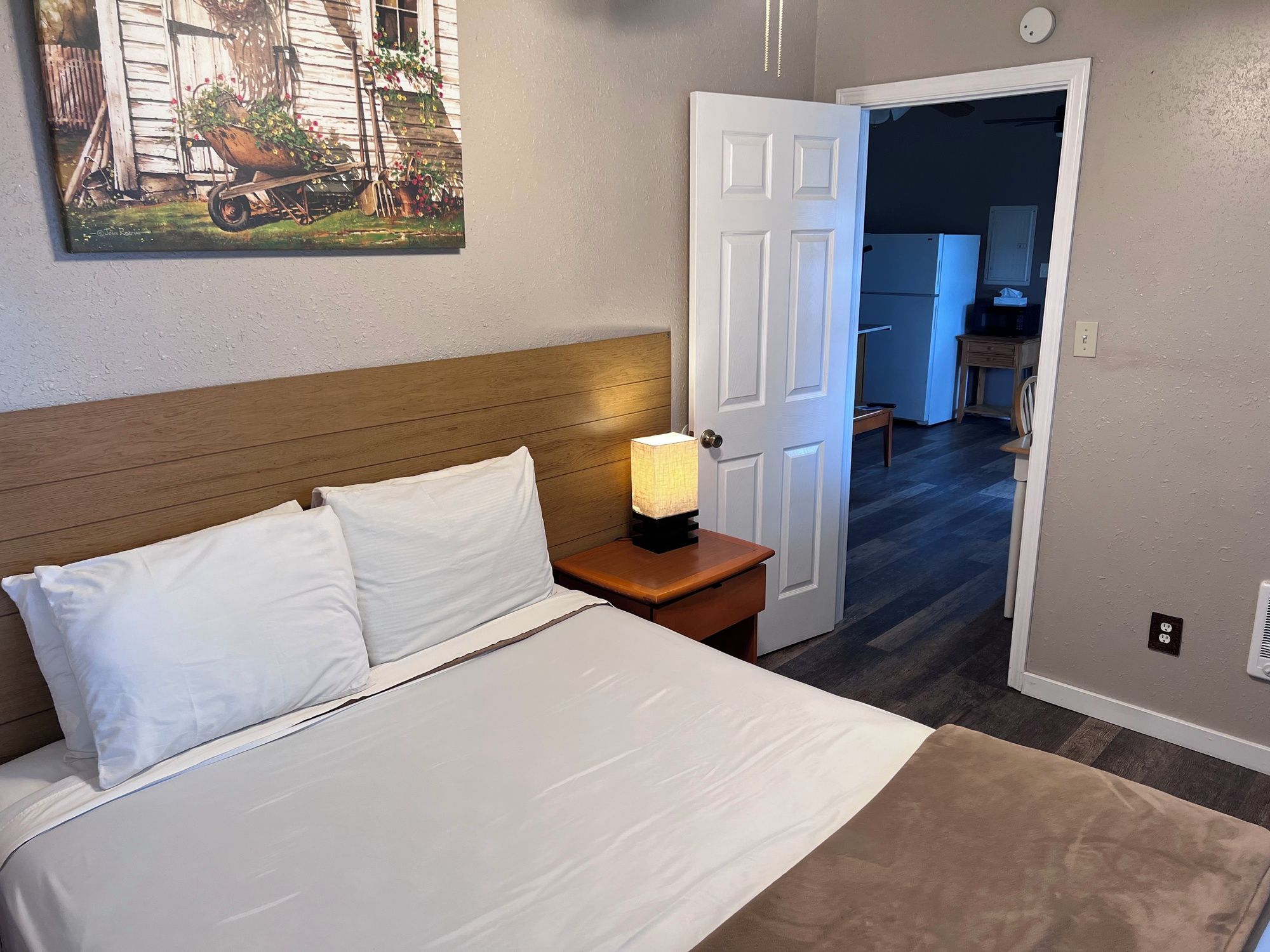A bedroom with a bed, a nightstand with a lamp, a door leading to a hallway, and a painting on the wall.