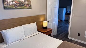 A bedroom with a bed, a nightstand with a lamp, a door leading to a hallway, and a painting on the wall.