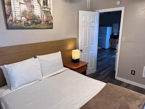 Quarto com cama, mesa de cabeceira com abajur, porta para um corredor e um quadro na parede.