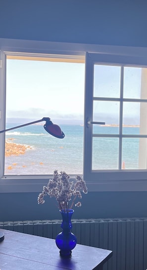 Ein Zimmer mit Fensterblick auf einen Strand, eine blaue Vase mit Trockenblumen auf einem Tisch und eine Schreibtischlampe.