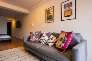 Ein modernes Wohnzimmer mit einem grauen Sofa, farbenfrohen Kissen, einem gerahmten Poster einer Straßenbahn und einem Bett im Hintergrund.