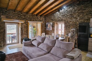 A cozy living room with a stone wall, wooden ceiling beams, a grey sofa, and a dining area with a table and chairs.