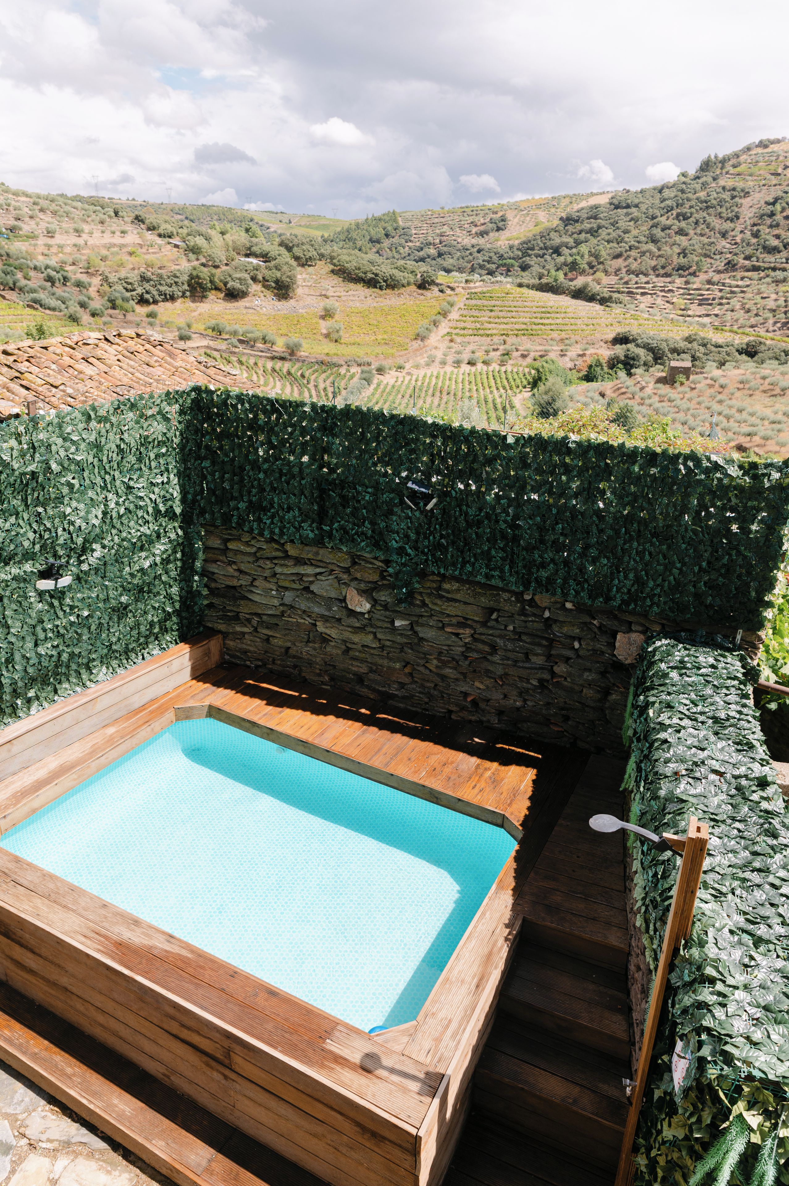 A hot tub with a stone wall and ivy-covered walls, surrounded by a wooden deck and a view of a vineyard.