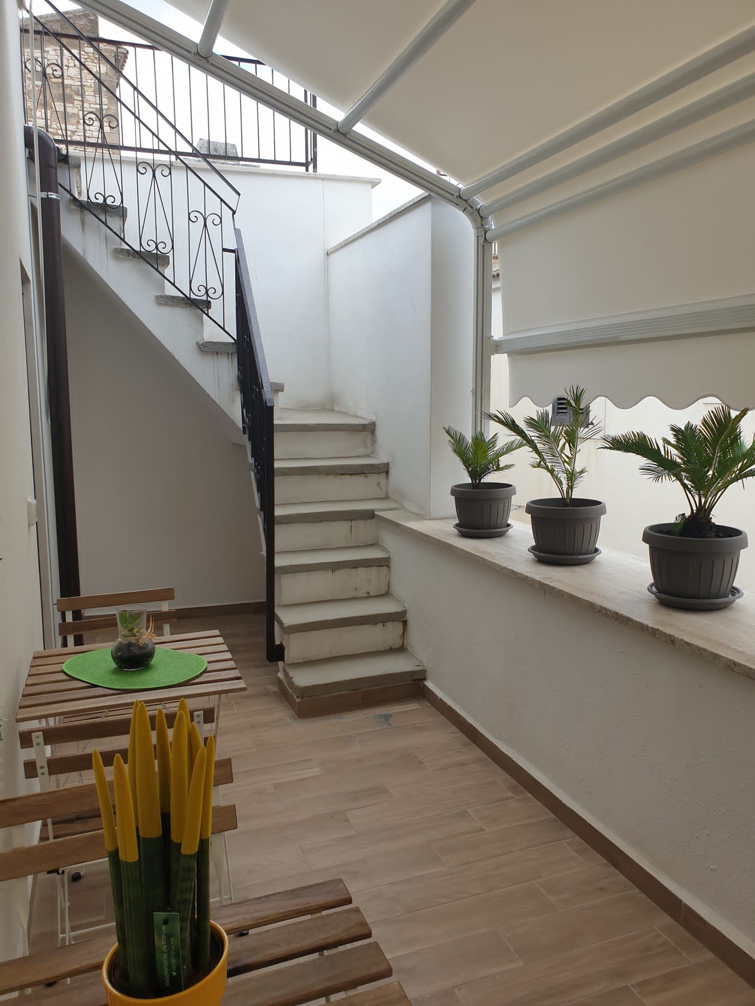 Escadaria que dá acesso a um andar superior, área de refeições com mesa e cadeiras, e plantas em vasos sobre um parapeito.
