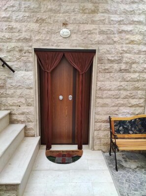 Uma porta de madeira com uma placa acima, um conjunto de degraus levando até a porta e um banco à direita.
