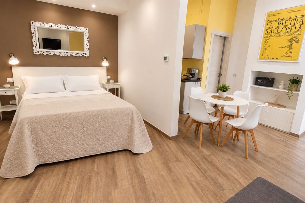 A hotel room with a bed, bedside tables, a desk with chairs, a small kitchenette, and a yellow wall with a poster.