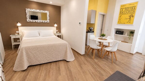 A hotel room with a bed, bedside tables, a desk with chairs, a small kitchenette, and a yellow wall with a poster.