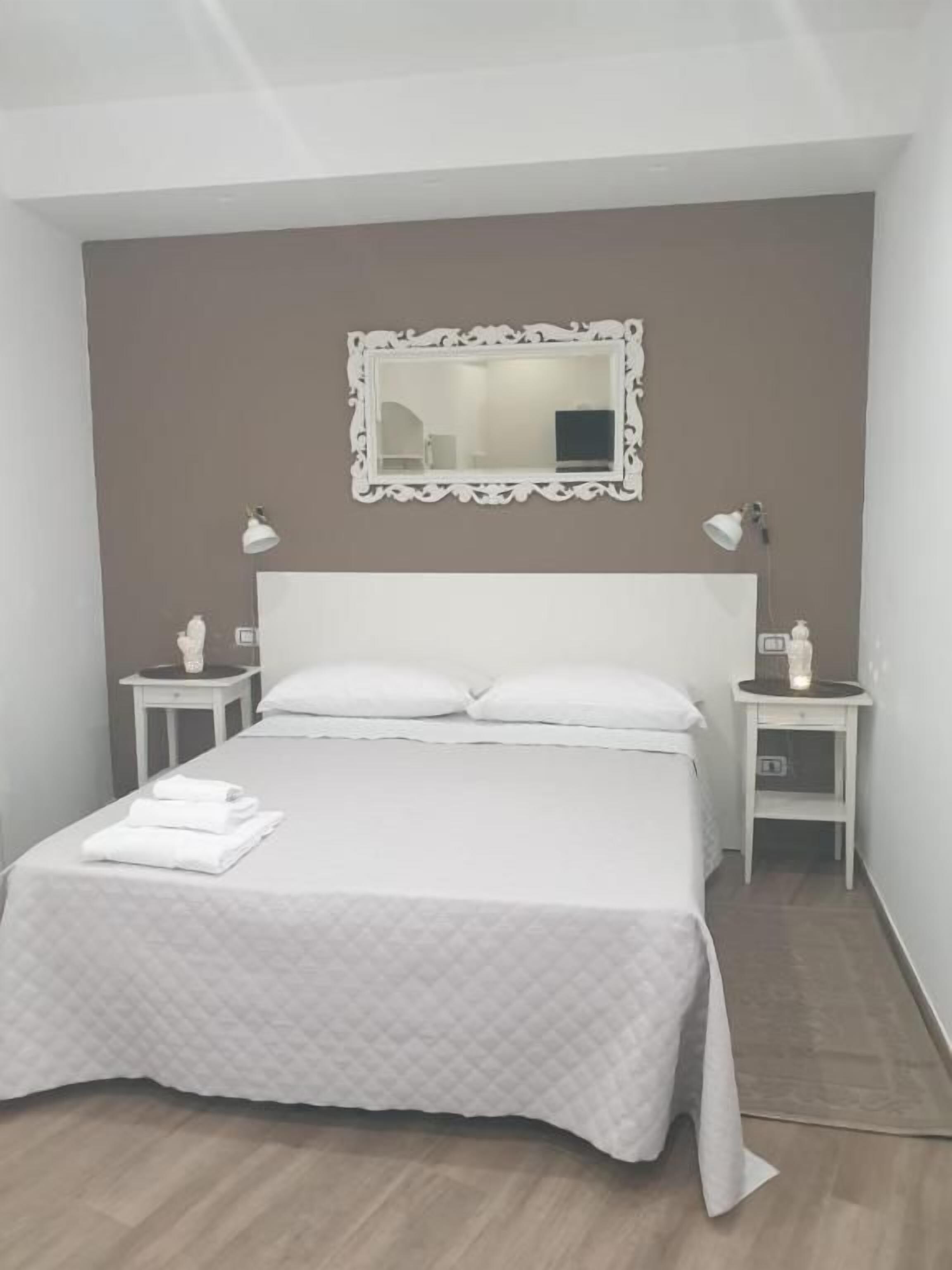 Una cama bien hecha con edredón y almohadas blancos, cabecera blanca, dos mesitas de noche con lámparas y un espejo en la pared.