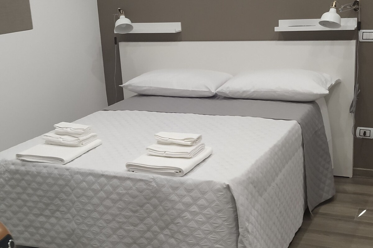 Una cama bien hecha con toallas dobladas, una mesita de noche y una lámpara de pared.