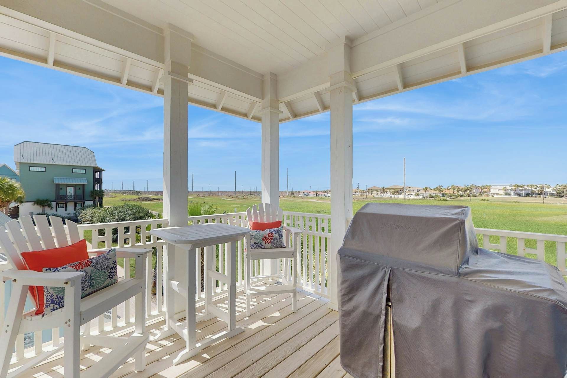 A covered outdoor seating area with white furniture, a covered grill, and a view of a residential area and beach.