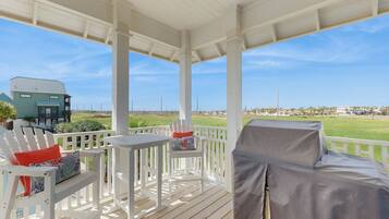 A covered outdoor seating area with white furniture, a covered grill, and a view of a residential area and beach.