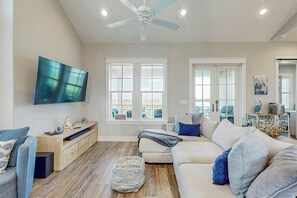 Ein geräumiges Wohnzimmer mit Deckenventilator, großen Fenstern, einem Flachbildfernseher und einer gemütlichen Sitzecke.
