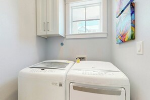 A laundry room with two white front-loading washing machines, a window, and a colorful abstract painting on the wall. - Palmilla Beach Resort-104 Agave Lane (Port Aransas)