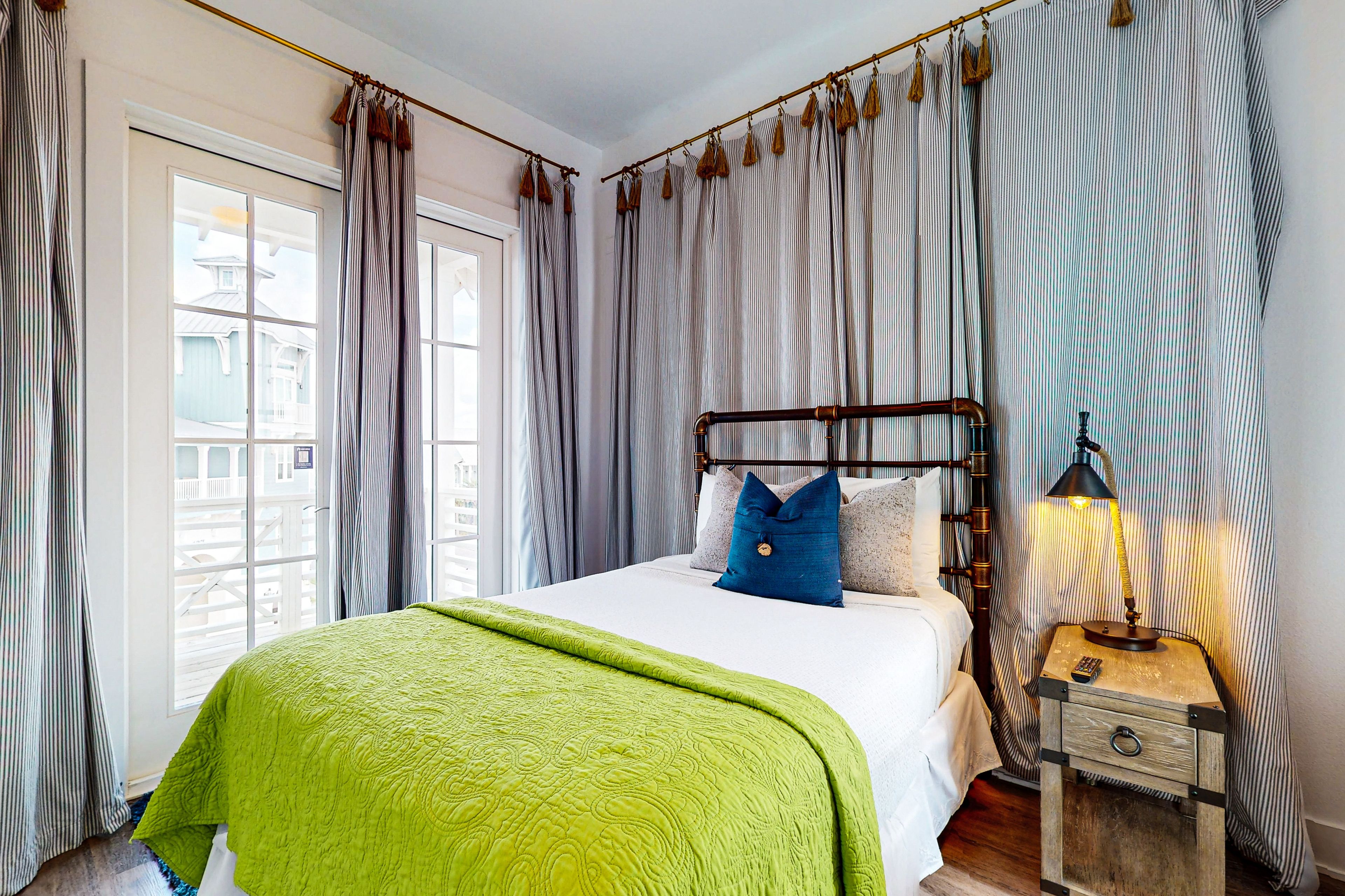 A neatly made bed with a green blanket, a wooden nightstand with a lamp, and striped curtains.