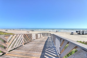 Een houten looppad dat naar een strand leidt, met voertuigen en een helderblauwe lucht.
