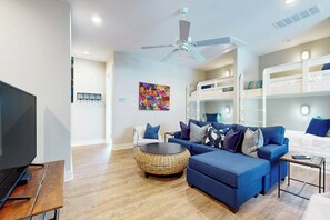 Een moderne woonkamer met een blauwe bank, een rond rieten salontafeltje, een flatscreen televisie en een plafondventilator.