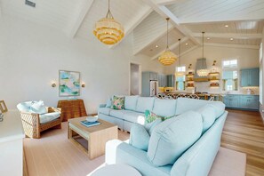 A spacious living room with a light blue sofa, wooden coffee table, and a wicker side table. A kitchen with light blue cabinets and a hanging light fixture is visible in the background. - Palmilla Beach Resort-135 Wild Indigo Street (Port Aransas)