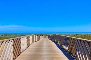 Ein hölzerner Steg führt zum Strand, der bewölktere Himmel ist blau.