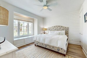 Ein Schlafzimmer mit einem Bett, Deckenventilator, Fenster mit Jalousien und einem Nachttisch mit Lampe.