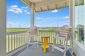 Eine überdachte Veranda mit zwei Adirondack-Stühlen und einem kleinen runden Tisch mit Blick auf einen Golfplatz.
