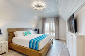 Ein Schlafzimmer mit einem hölzernen Bett, weißer Bettwäsche, einer blau-weißen Decke, einer Deckenleuchte, einem Fernseher und einem Fenster mit Vorhängen.