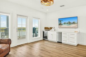 A spacious room with a wooden floor, a brown leather sofa, a kitchen area with white cabinets, and a large window overlooking a deck. - Palmilla Beach Resort-152 Sunrise Avenue (Port Aransas)