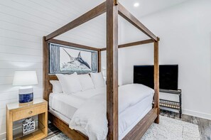 Ein Bett mit Baldachin, ausgestattet mit weißer Bettwäsche, ein gerahmtes Kunstwerk darüber, ein Nachttisch mit Lampe sowie ein Fernseher auf einem Sockel.
