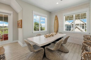 A dining area with a white table and wicker chairs, a surfboard on the wall, and a view of a building complex. - Palmilla Beach Resort-158 Sunset Avenue (Port Aransas)