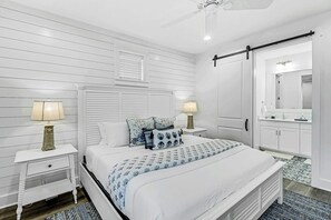 Ein Schlafzimmer mit einem Bett, zwei Nachttischen, einem Schminktisch, einem Deckenventilator, einer Tür und einem Badezimmer mit Spiegel und Waschbecken.