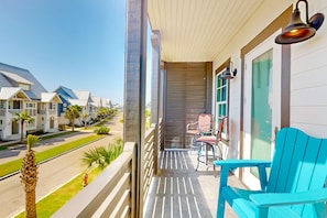 A balcony with a view of a street lined with houses, a set of chairs, and a door with a sliding shutter. - Palmilla Beach Resort-169 Spanish Dagger Lane (Port Aransas)