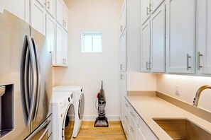 Een smalle keuken met witte kasten, een roestvrijstalen koelkast, een wasmachine en een stofzuiger.