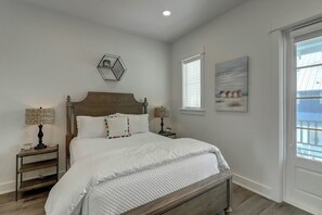 Ein Schlafzimmer mit einem hölzernen Bett, zwei Nachttischen mit Lampen, einer geometrischen Wanddekoration, einem Gemälde zum Thema Strand und einem Fenster mit Jalousien.