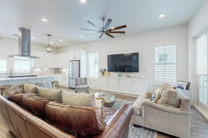 A modern living room with a brown leather sofa, a beige armchair, and a dining area with white cabinetry and a large flat-screen TV. - Palmilla Beach Resort-190 Sage Lane (Port Aransas)