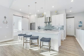 A modern kitchen with white cabinetry, a central island with bar stools, and a stainless steel refrigerator. - Palmilla Beach Resort-200 Sunset Avenue (Port Aransas)