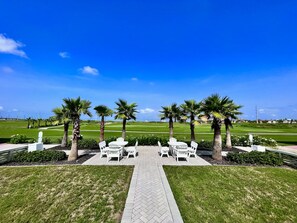A golf course with a paved walkway, white chairs, and palm trees. - Palmilla Beach Resort-204 Sunset Avenue (Port Aransas)