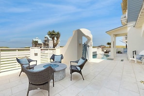 An outdoor patio with a round table and chairs, a swimming pool, and a view of the ocean and buildings in the background. - Palmilla Beach Resort-218 Sunrise Avenue (Port Aransas)