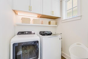 A laundry room with two washing machines, a shelf with baskets, and a window with blinds. - Palmilla Beach Resort-220 Sunrise Avenue (Port Aransas)
